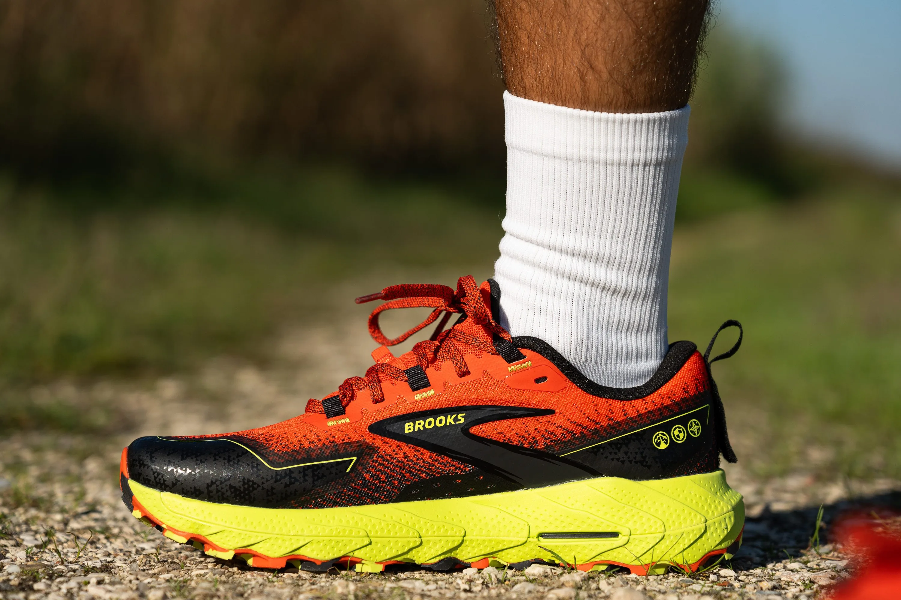 A Brooks Cascadia trail shoe on a wet dirt trail with small rocks, highlighting a structured upper and moderate lugs