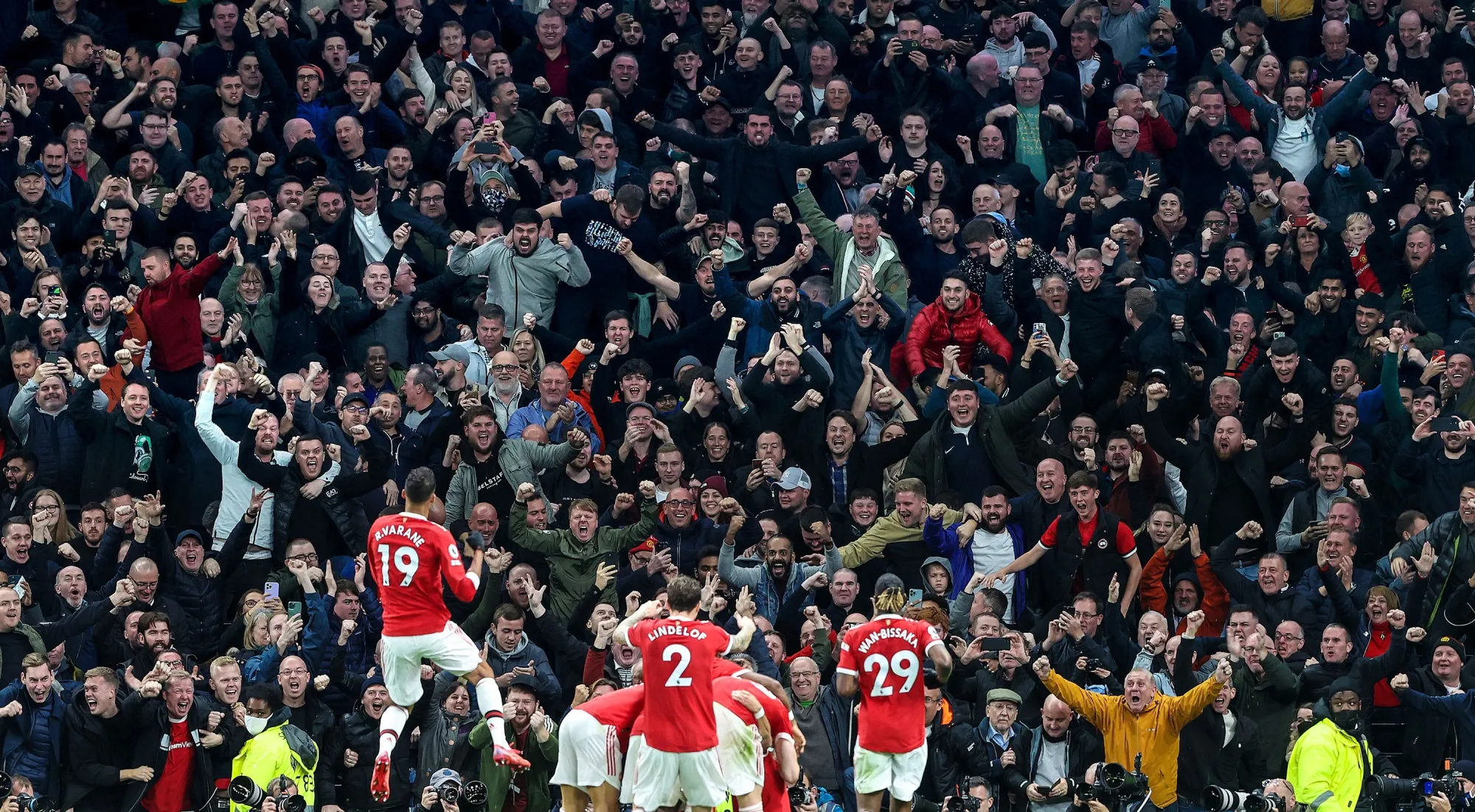 A Premier League stadium crowd during a night match, fans holding scarves aloft as the teams line up on the pitch
