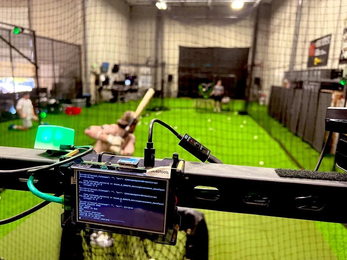 A baseball pitcher throwing in an indoor training facility while coaches monitor radar and ball-tracking equipment behind the mound
