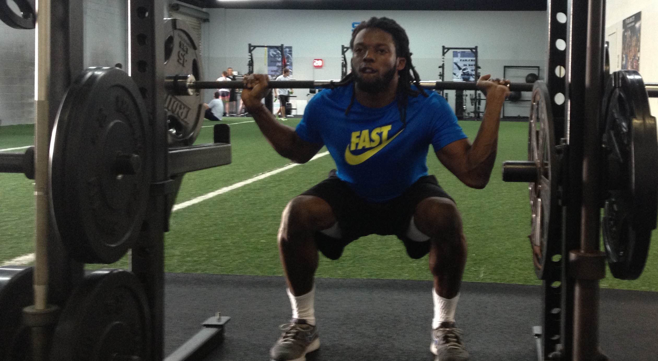 A basketball athlete in a weight room performing a back squat under a barbell with a coach spotting from behind