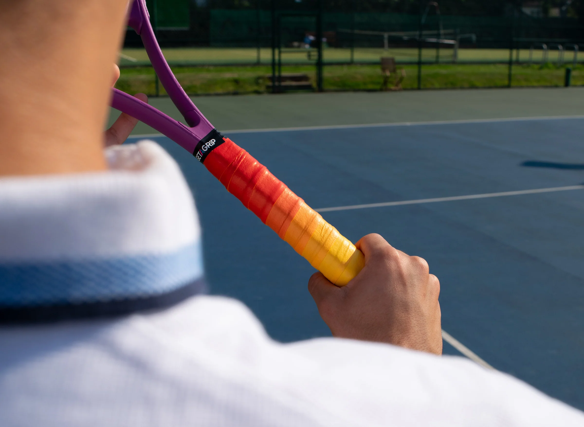 A close-up photo of a tennis racket handle with a fresh overgrip being wrapped by a player sitting on a court bench, with tennis balls nearby