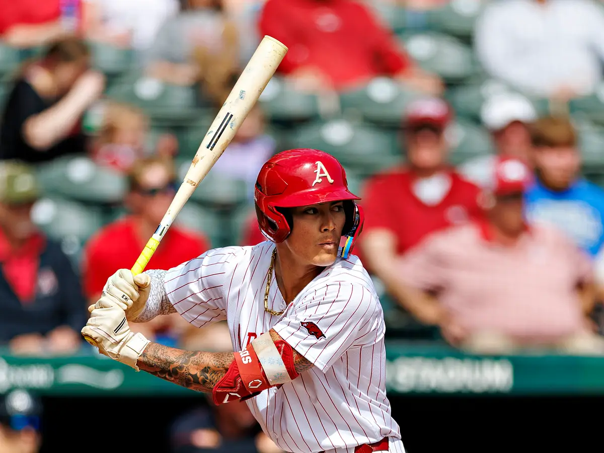 A college baseball hitter listed as the designated hitter taking an at-bat in a packed stadium, with the dugout and fans visible behind home plate