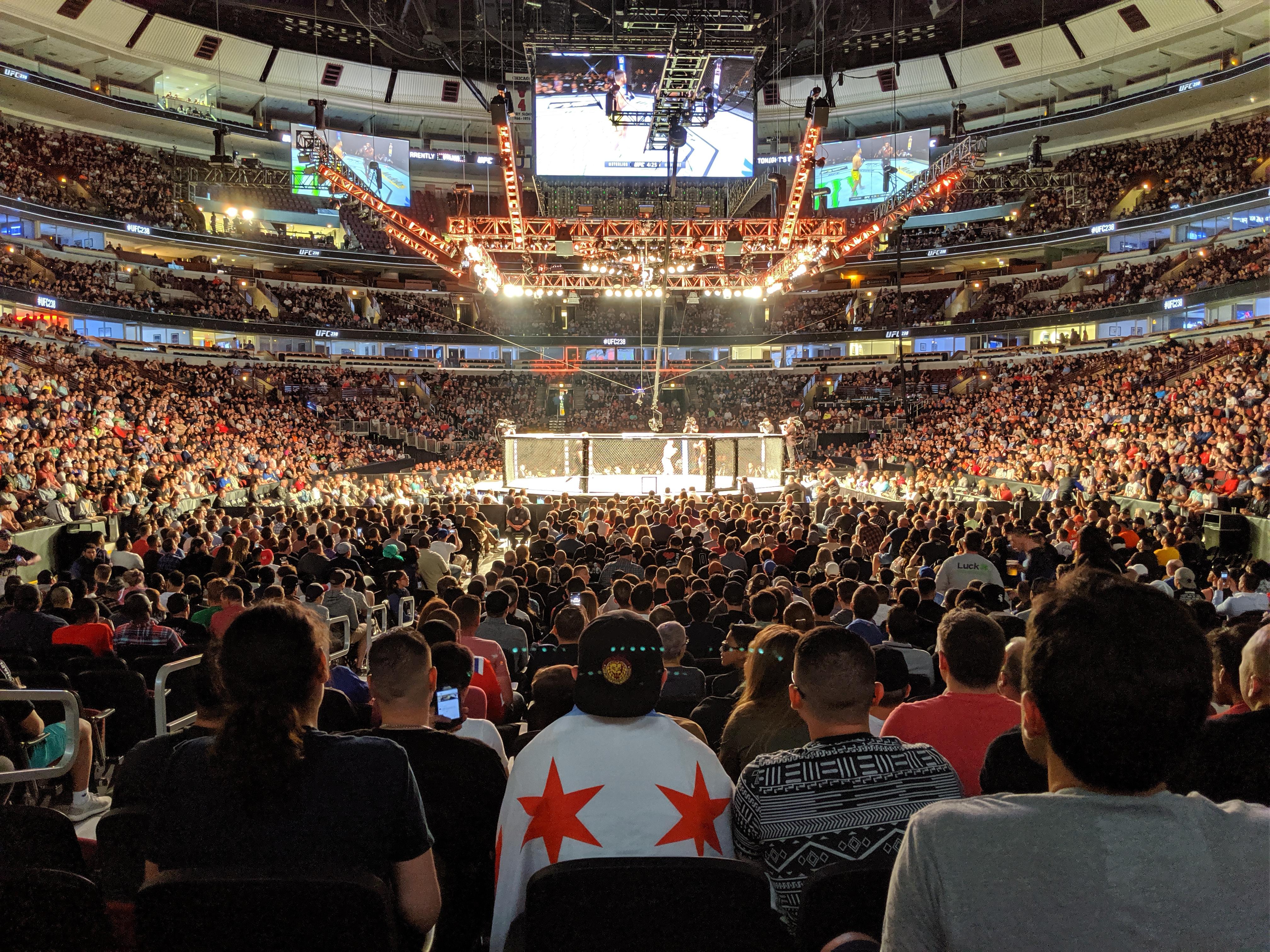 A crowd in an arena watching a UFC fight as the fighters stand in the octagon awaiting the judges' decision