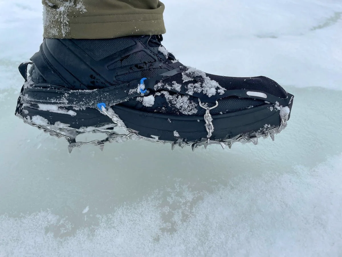A hiker wearing microspikes over hiking boots while walking on a packed snowy trail in a forest, mid-stride action shot