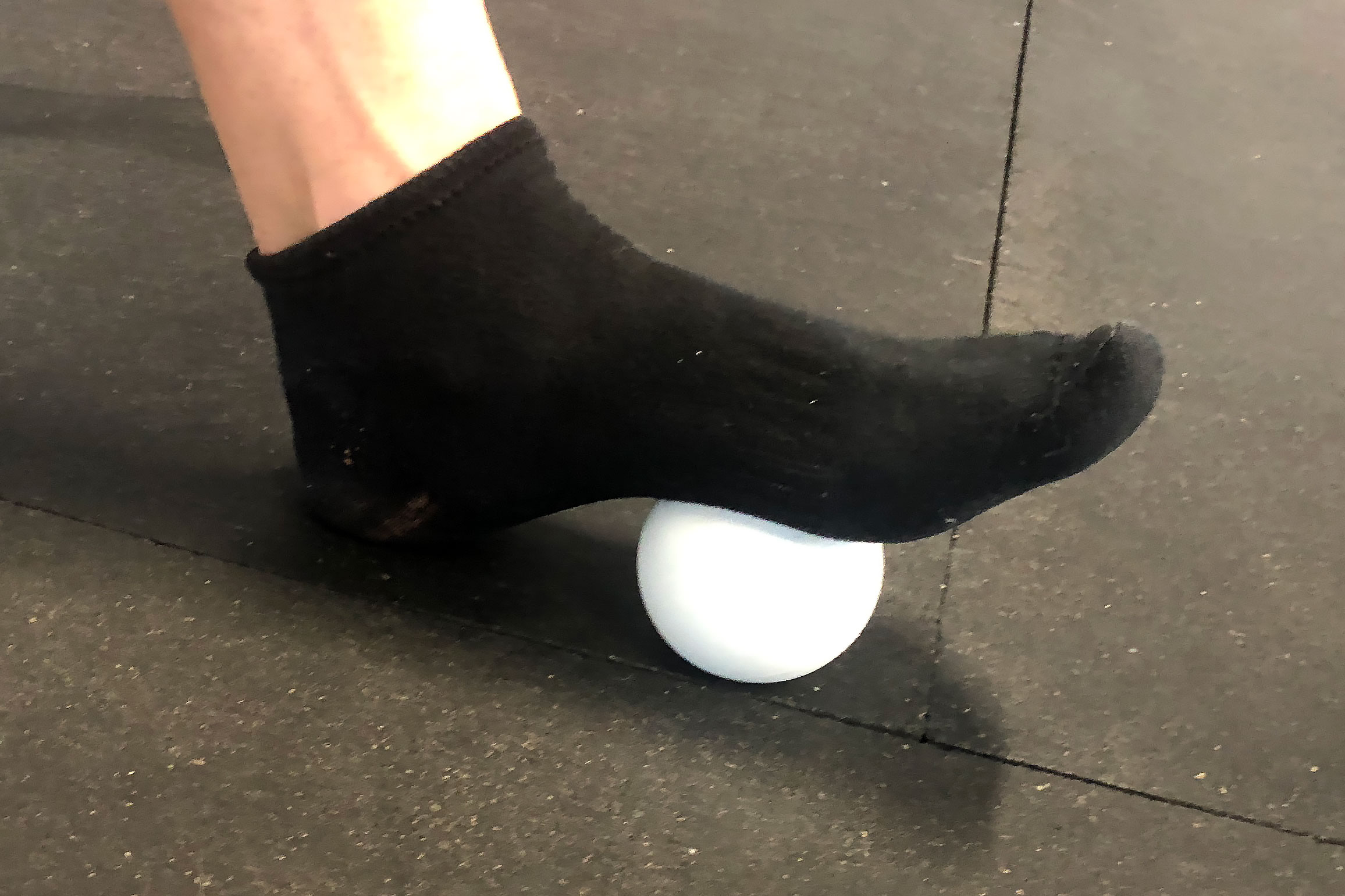 A lacrosse ball on a gym floor next to a runner’s shoe and sock, set up for a post-run foot rolling session.