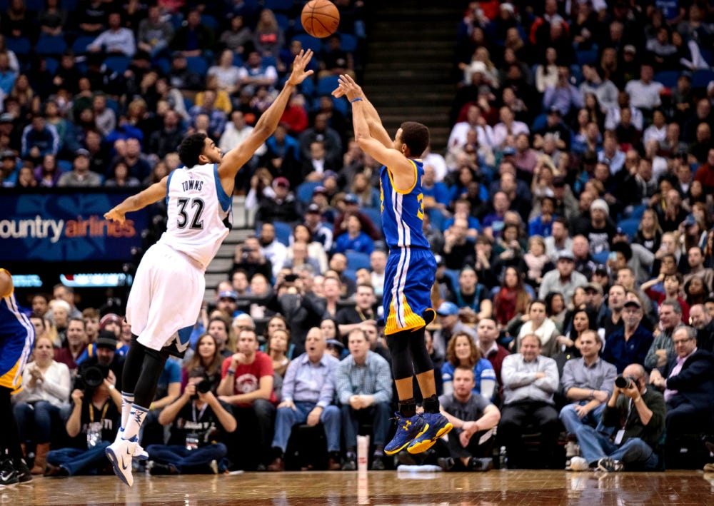 A professional basketball defender contesting a jump shot with an outstretched arm as the shooter releases the ball near the top of the key