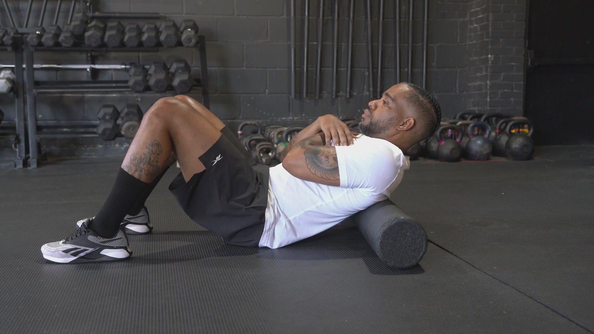 A professional basketball player on a hardwood court using a foam roller on the hamstrings during warmup, with teammates and practice gear nearby