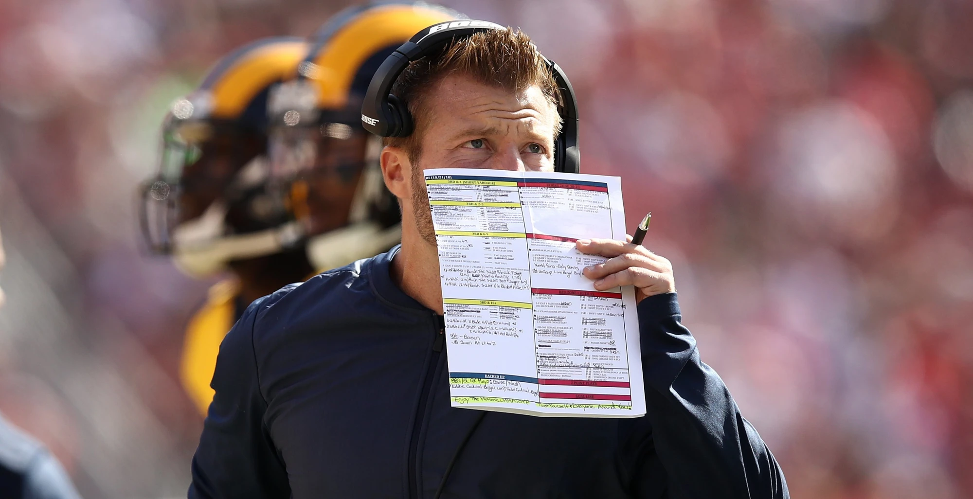 A professional football head coach on the sideline holding a play sheet and speaking into a headset during a close game