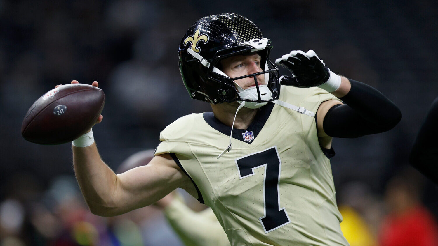 A professional football quarterback in uniform stepping into a throw while a defender closes in, stadium lights and crowd visible in the background