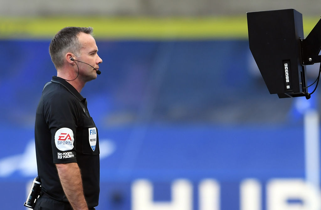 A referee standing at a pitch-side review screen with one hand to an earpiece while players from both teams wait near midfield during a night match
