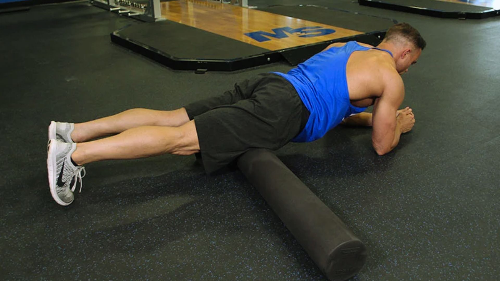 A runner foam rolling their quadriceps on a black mat in a gym, water bottle and shoes nearby