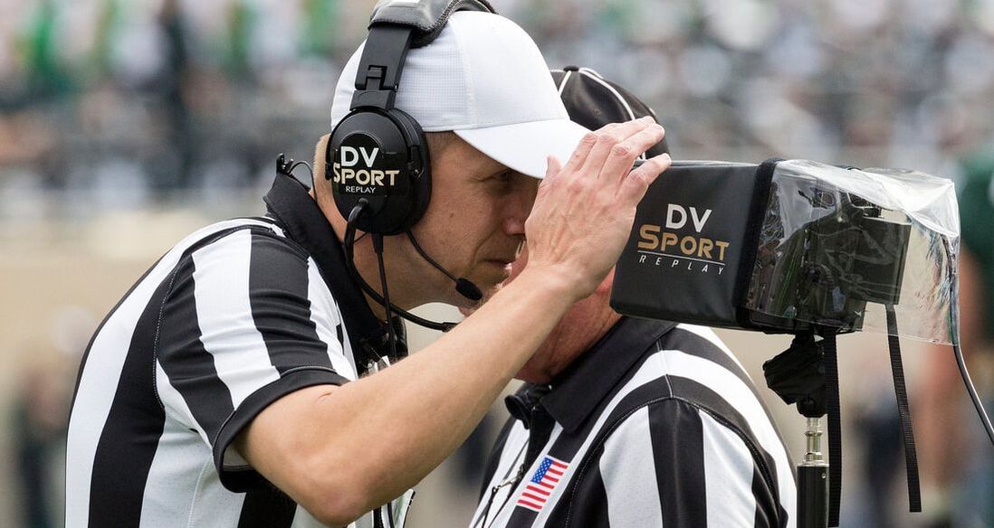 A sideline television camera crew filming an American football game while a replay operator monitors screens near the field, live sports production atmosphere