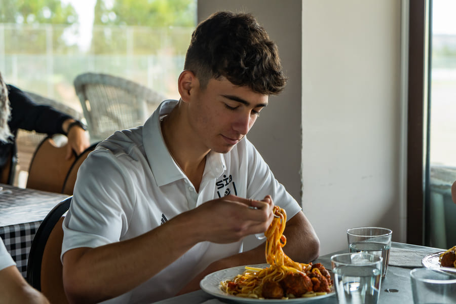 A soccer player in training gear eating a plate of pasta with simple tomato sauce at a kitchen table