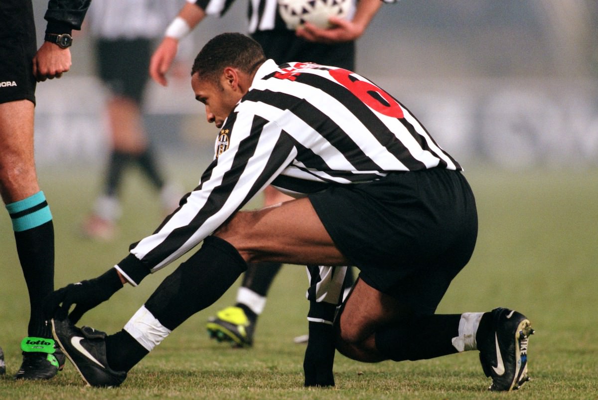 A soccer player sitting on the sideline holding their calf while a teammate offers support during a match