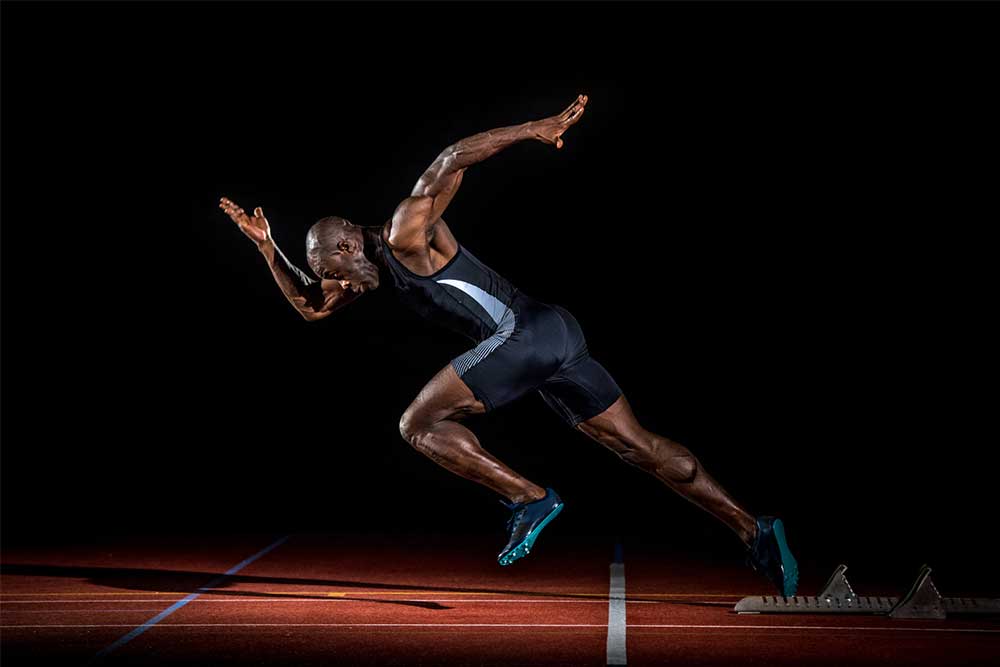 A sprinter captured from the side at top speed on a track, showing tall posture, strong front-side mechanics, and a foot striking close under the hips