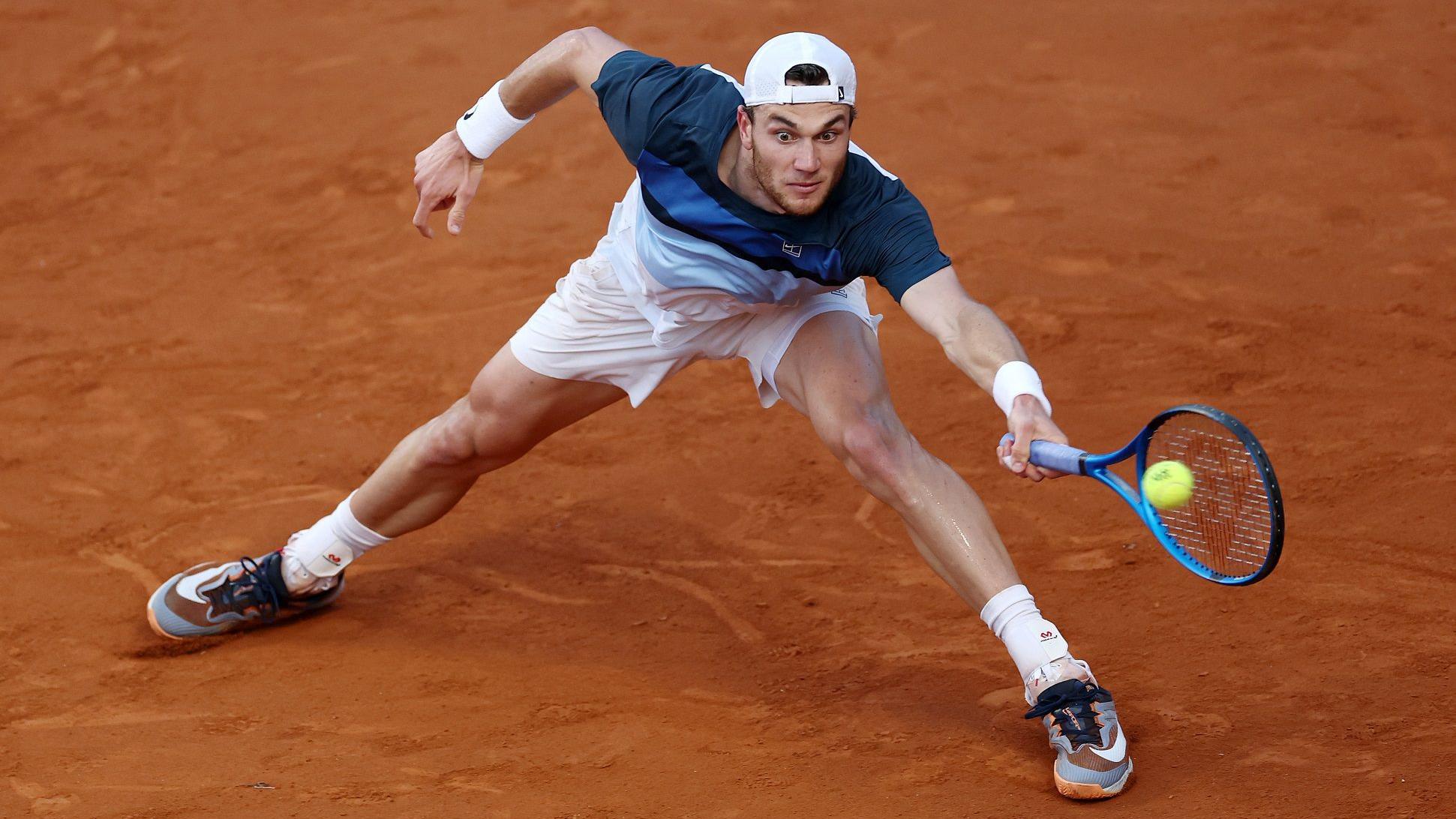 A tennis player hitting a heavy topspin forehand on a clay court, with the ball rising off the strings and dust kicked up near the baseline