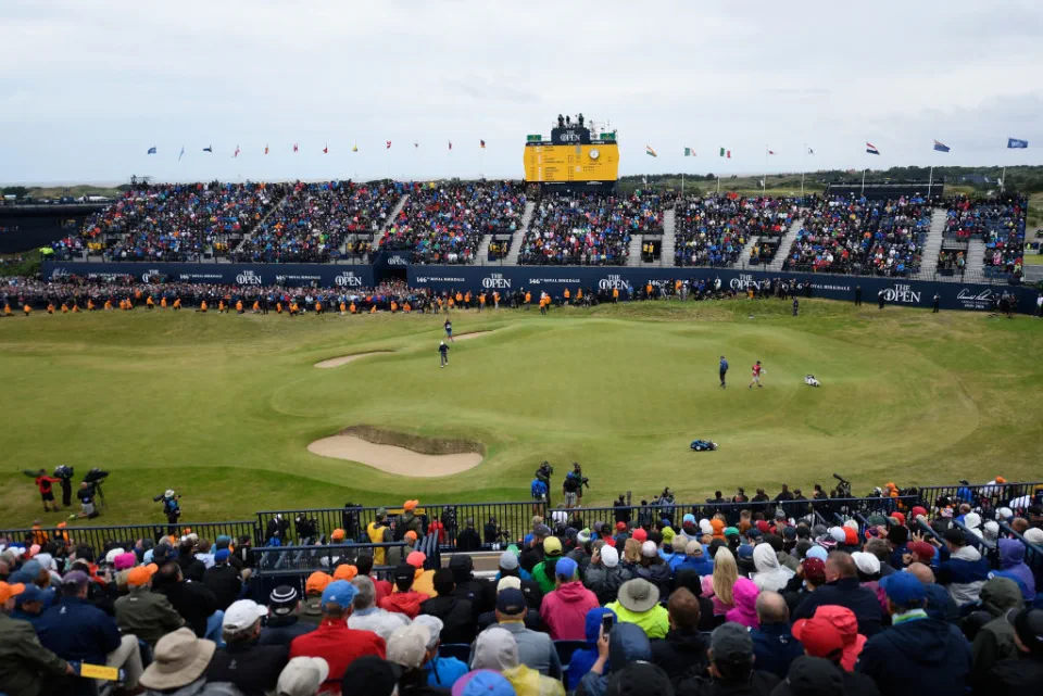 A wide view of Royal Birkdale during the 2026 Open Championship with grandstands surrounding a green and fans watching players approach in bright daylight
