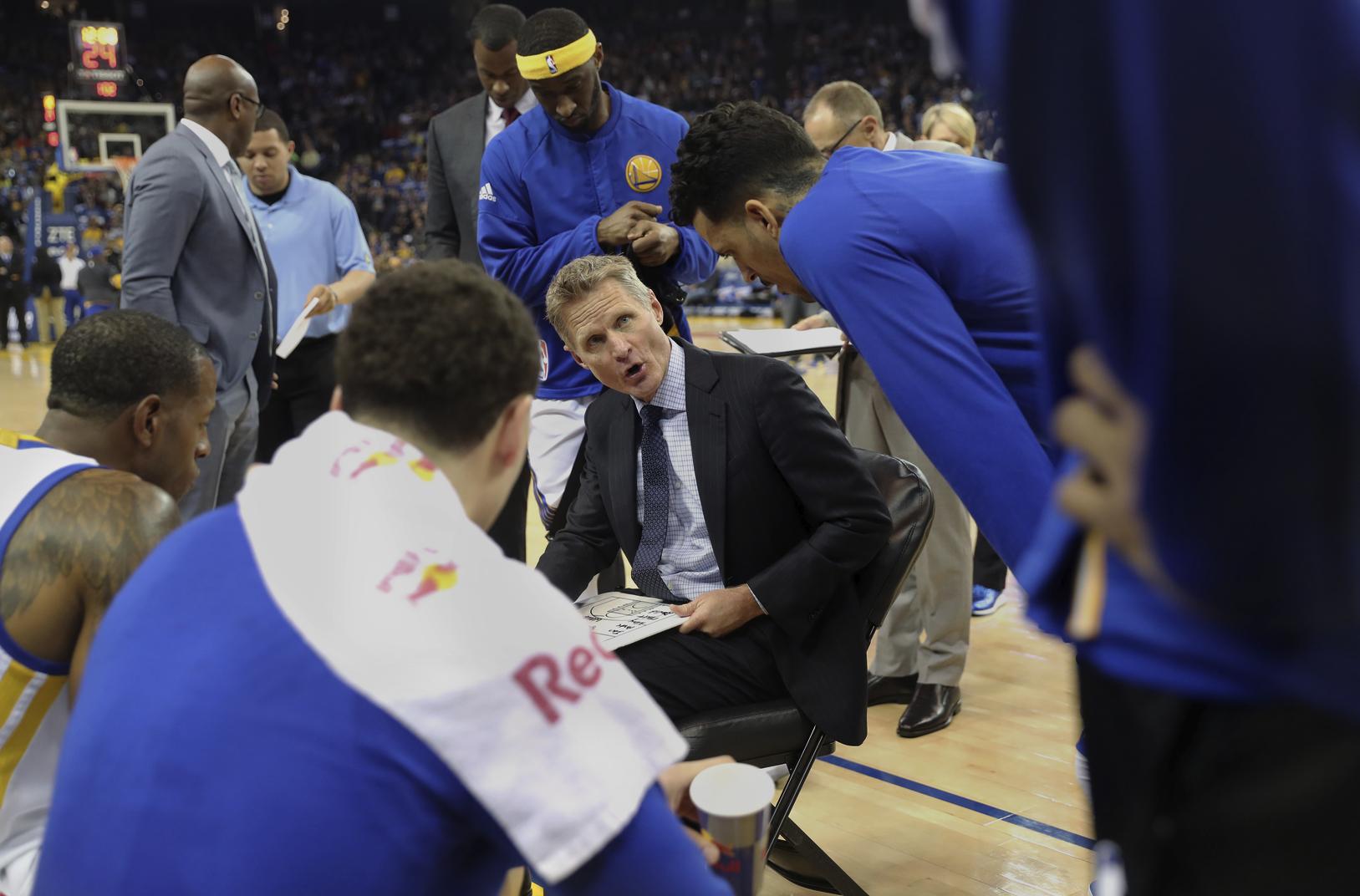 An NBA coach during a timeout drawing a play on a clipboard as players lean in and listen.