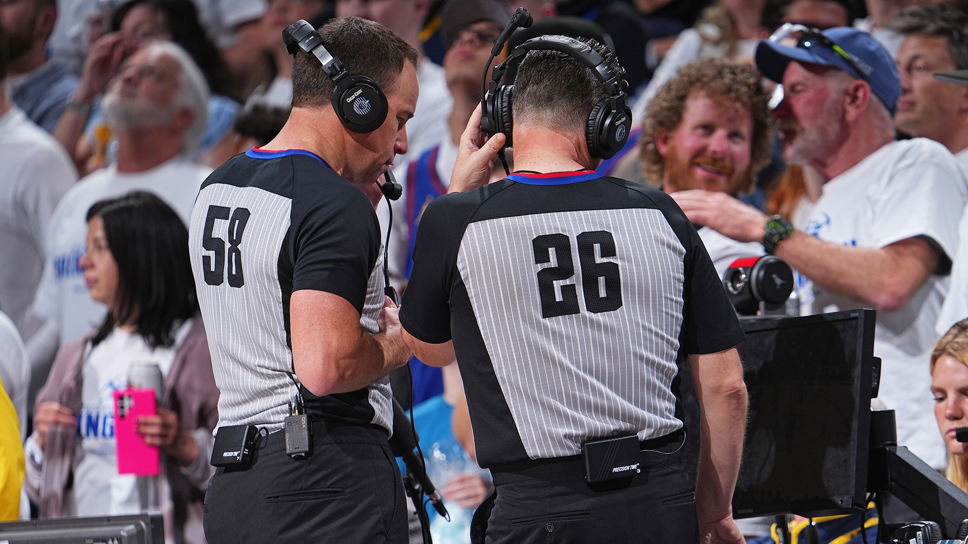 An NBA head coach on the sideline forming the challenge signal with both hands while referees confer near the scorer’s table