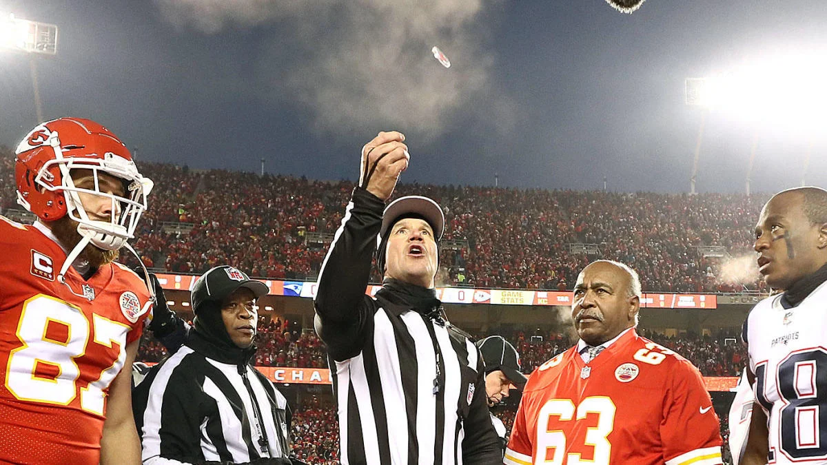 An NFL referee holding a coin at midfield during an overtime coin toss with two team captains standing nearby in a packed stadium