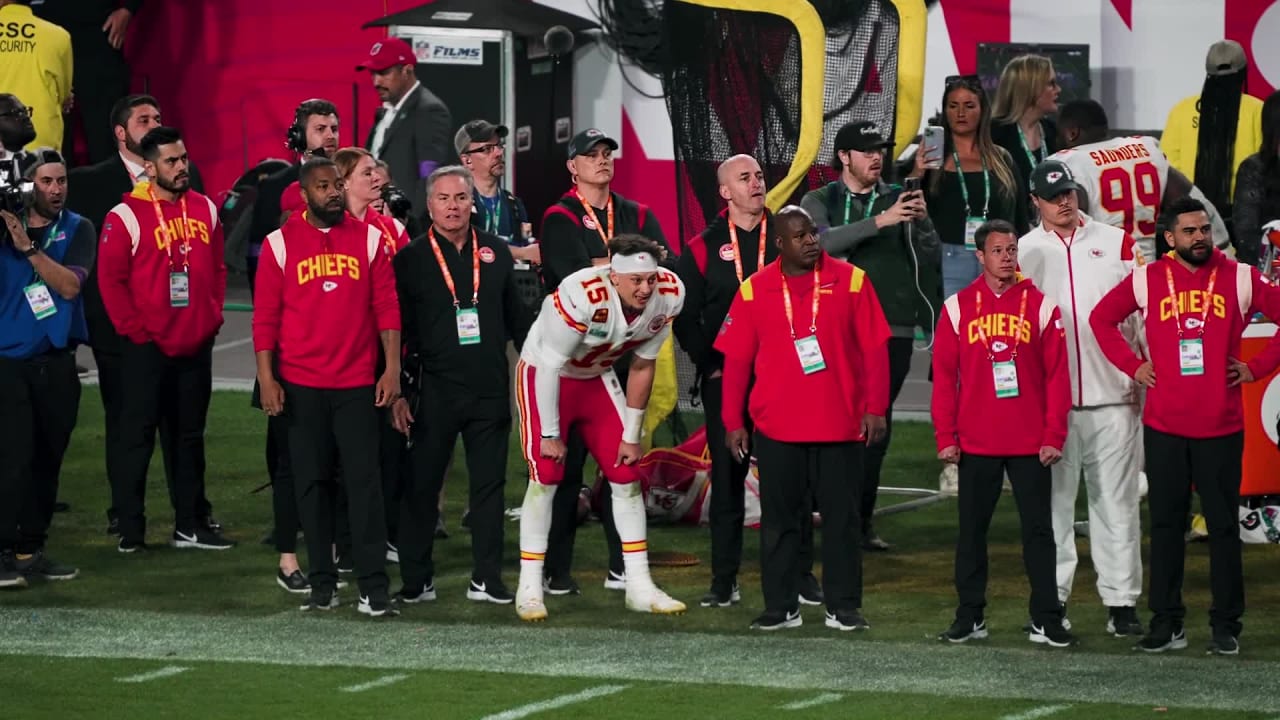An NFL sideline scene with coaches and players in Kansas City Chiefs gear during a 2024 regular-season game, with the stadium crowd blurred in the background