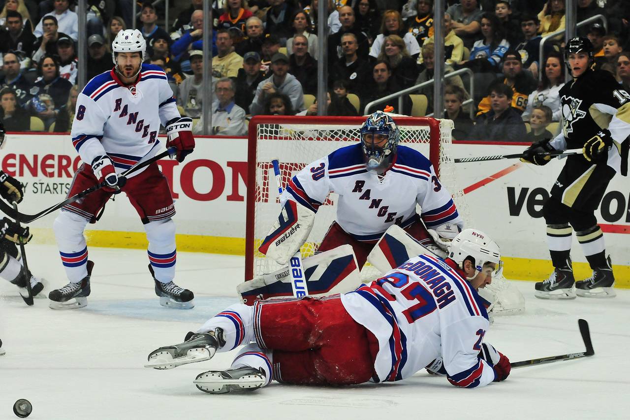 An NHL defenseman dropping to one knee to block a slap shot from the point, with the puck mid-flight and the goalie tracking behind him in the crease