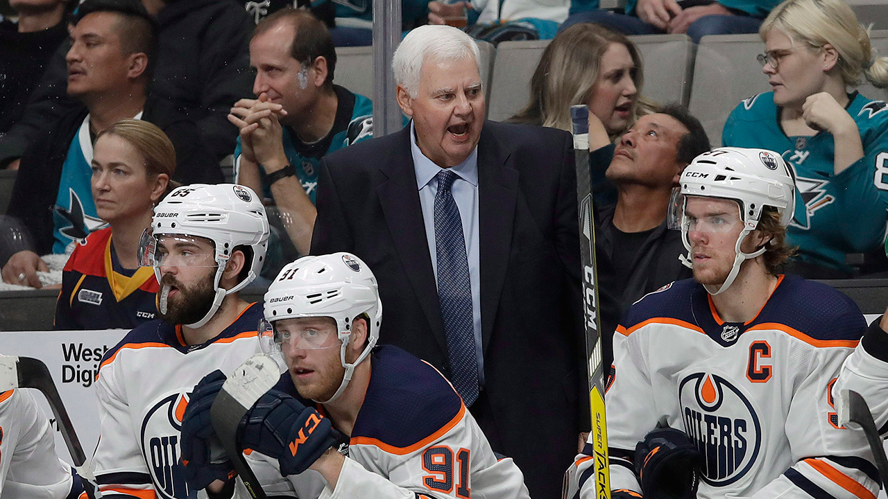 An NHL head coach standing behind the bench during a game, leaning forward with arms crossed while players sit in a row wearing helmets and gloves