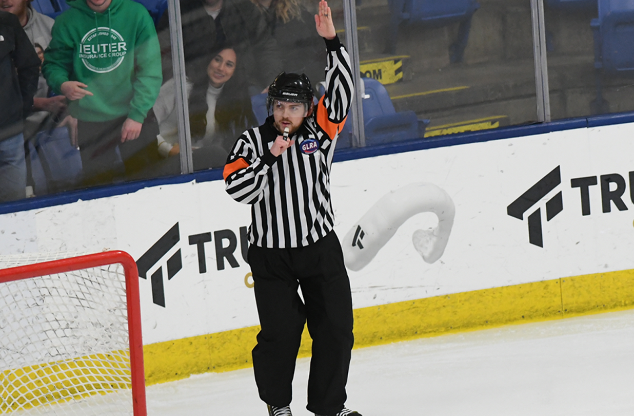 An NHL linesman with one arm raised to signal a delayed icing call as players skate toward the end boards during live play