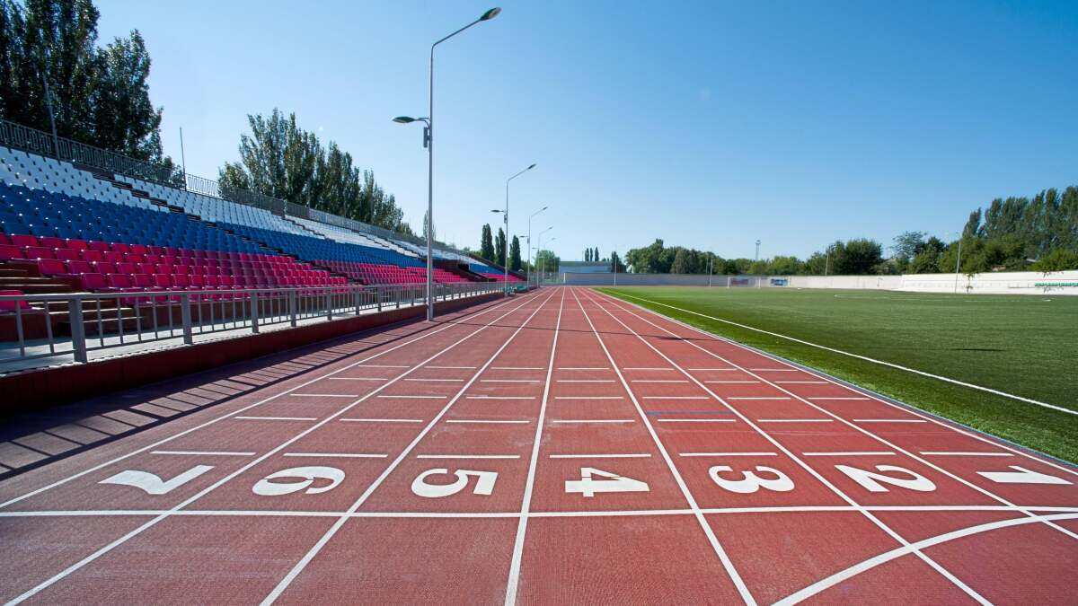 An empty outdoor rubber running track with lane lines curving past a football field on a clear morning