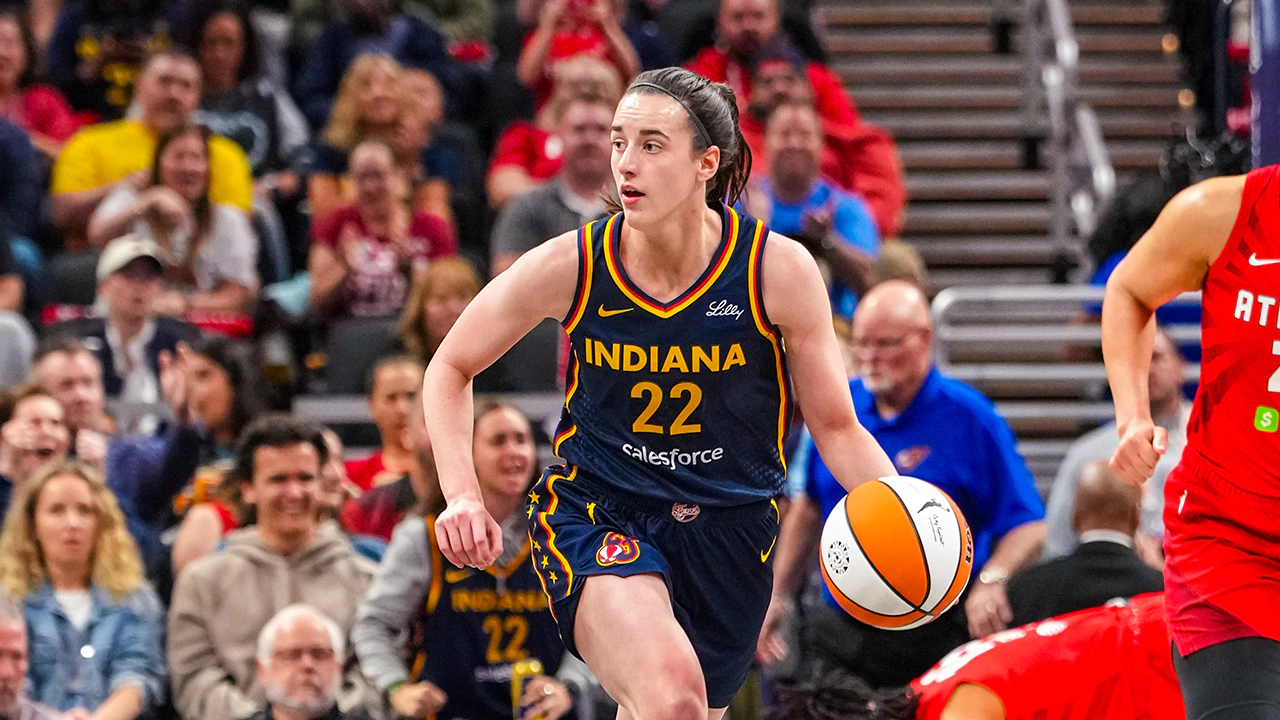 Caitlin Clark in an Indiana Fever uniform dribbling at the top of the key with a defender closing, eyes up reading the floor