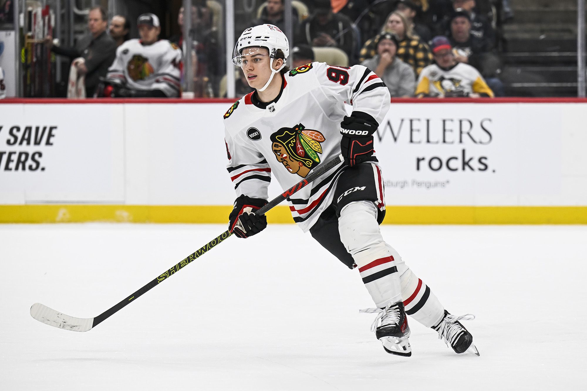 Connor Bedard in a Chicago Blackhawks jersey skating with the puck on his forehand, eyes up as a defender angles toward him