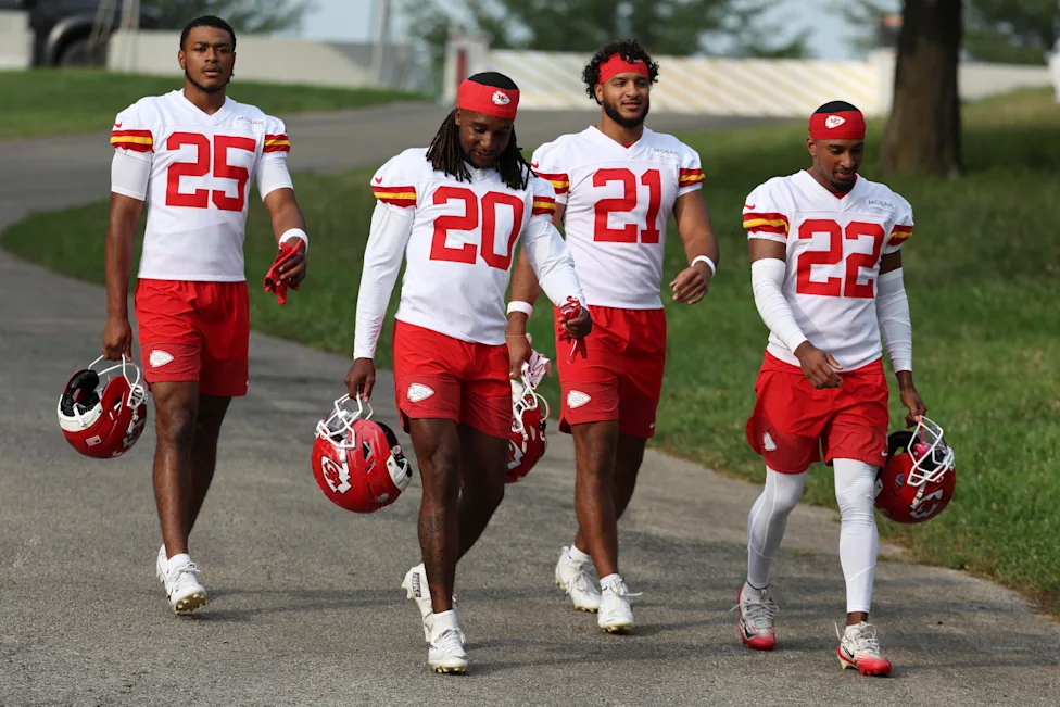 Defensive backs running a drill during NFL training camp while coaches watch from the sideline