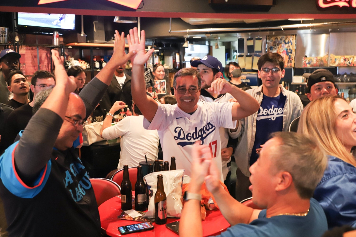 Fans in a neighborhood sports bar watching a live game on a large television, one person holding a phone showing a highlight clip, candid sports night atmosphere