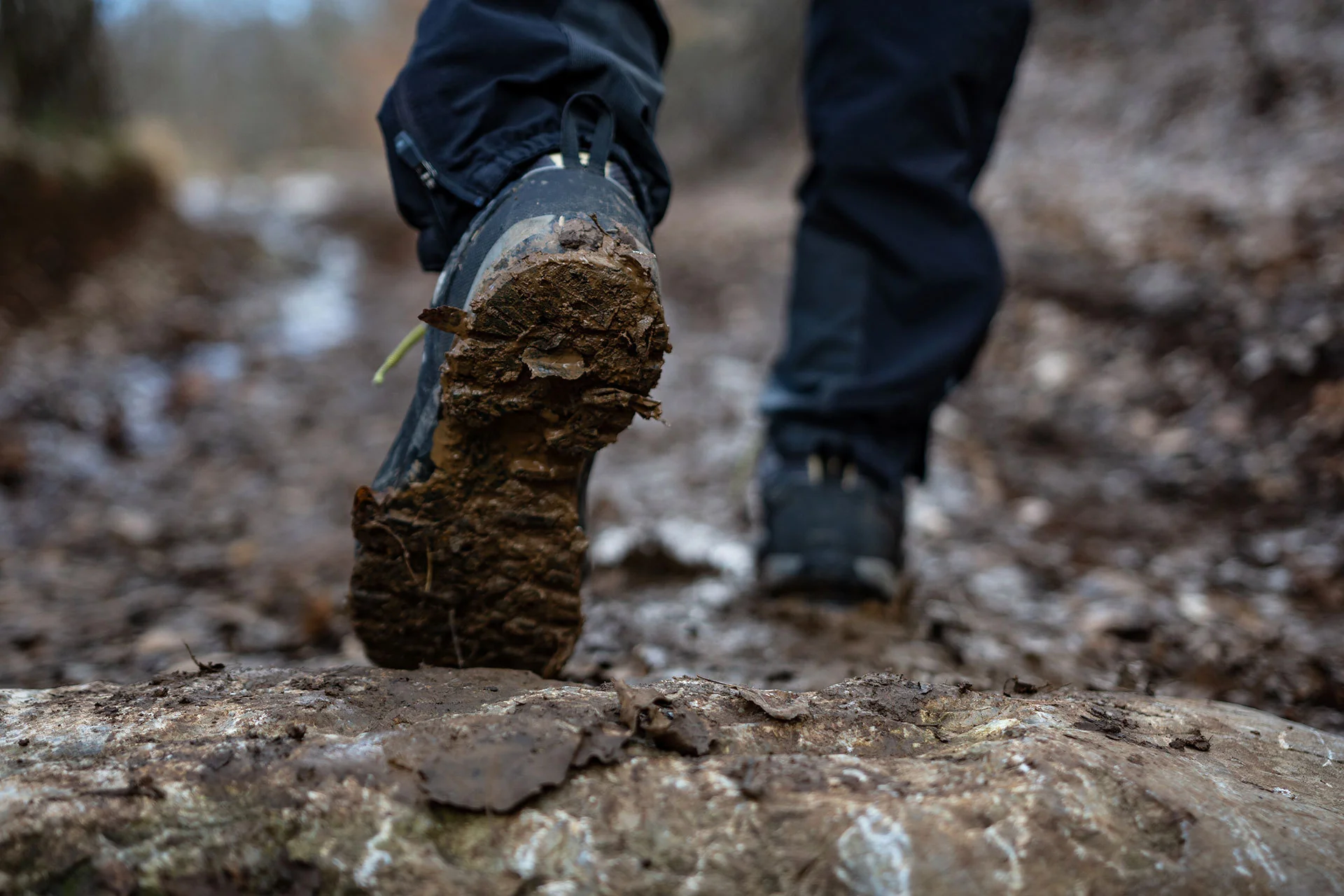 Hands using a soft brush to scrub dried mud from the seams of a hiking boot near a utility sink, close-up action shot