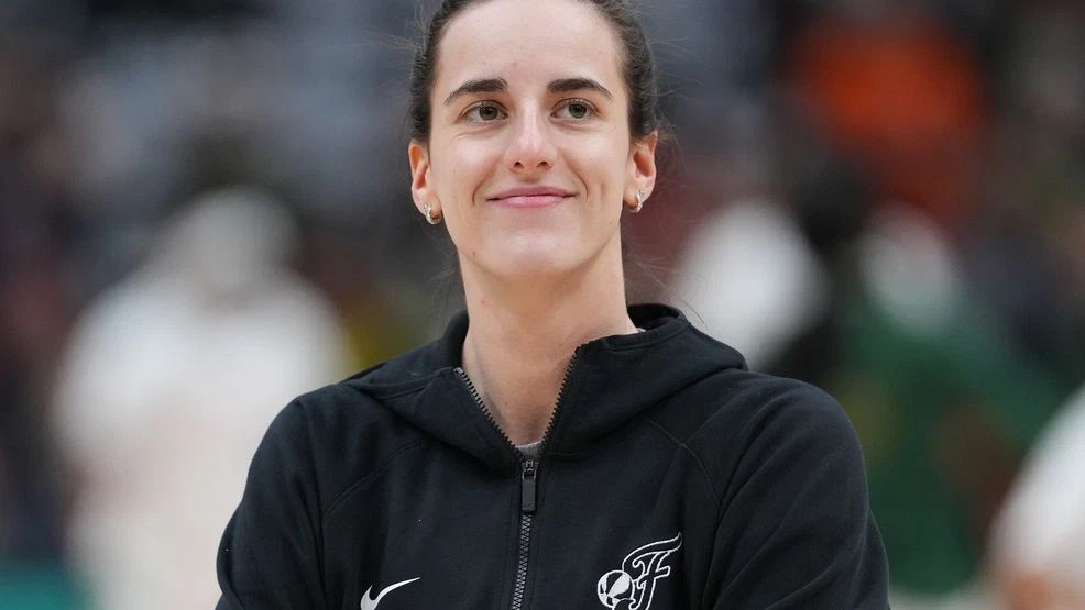 Indiana Fever guard Caitlin Clark on the bench during a game, watching the action with an intense expression while coaches and teammates stand nearby