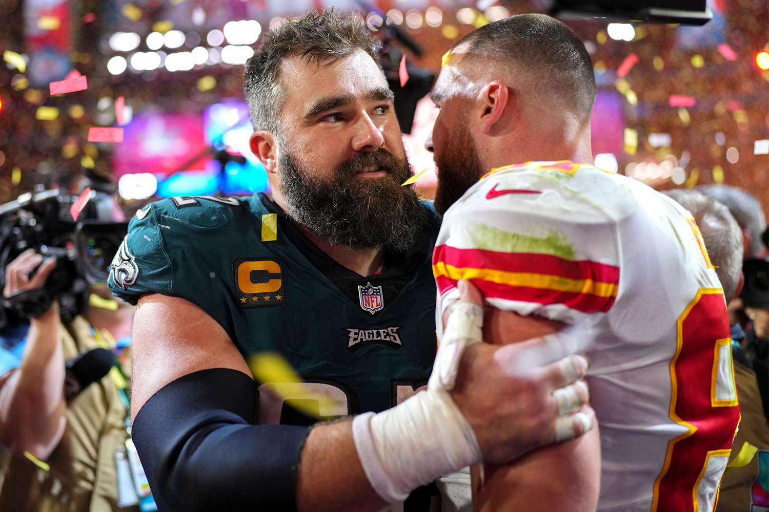 Jason Kelce and Travis Kelce on the field after the Super Bowl, surrounded by players and staff under bright stadium lights.