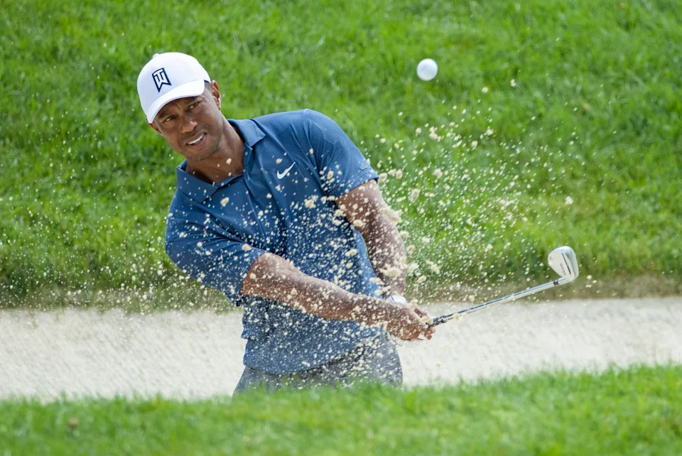 Tiger Woods walking along a fairway during a PGA Tour event, wearing golf attire and carrying a club, with fans watching from behind the ropes