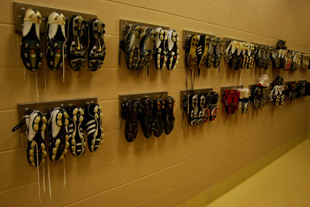 Two pairs of soccer shoes, one turf model with rubber nubs and one firm ground model with molded studs, placed on a wooden locker room bench before a game