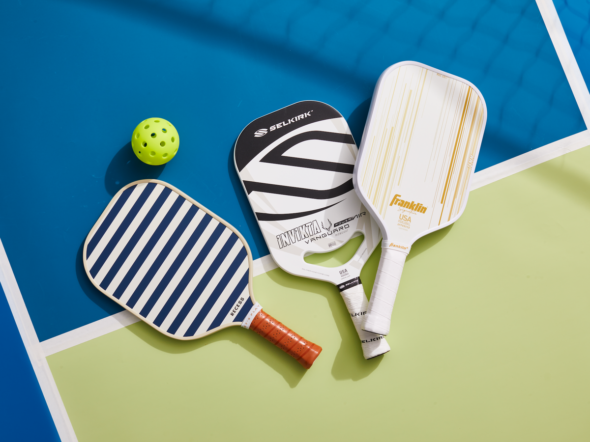 Two pickleball paddles resting courtside next to pickleballs, showing different paddle shapes and handle lengths