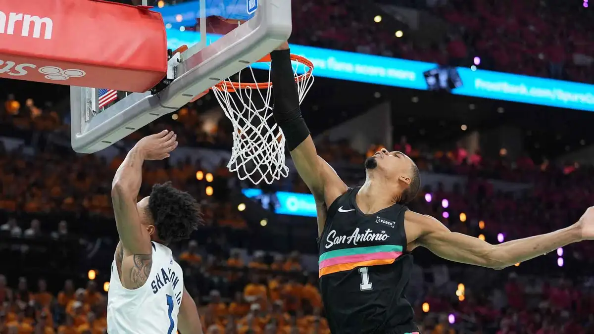 Victor Wembanyama leaping near the rim to block a shot, with the ball redirected above the cylinder as players look up