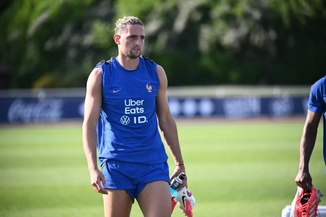 William Saliba in France training gear on a World Cup training pitch, walking with a focused expression as staff and teammates move in the background, news photography style