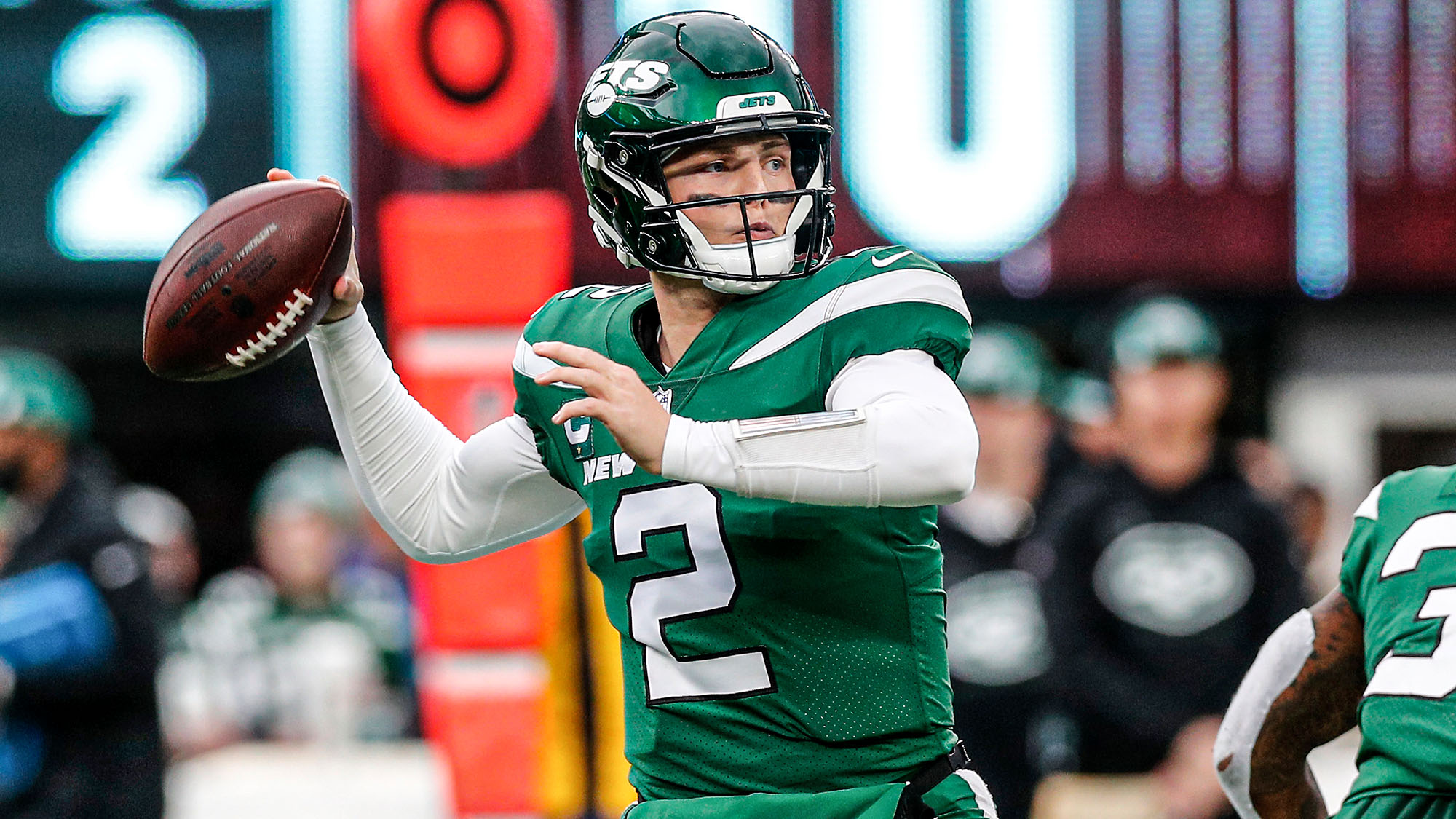 Zach Wilson in a New York Jets uniform warming up on the field before an NFL game, holding a football and looking toward teammates